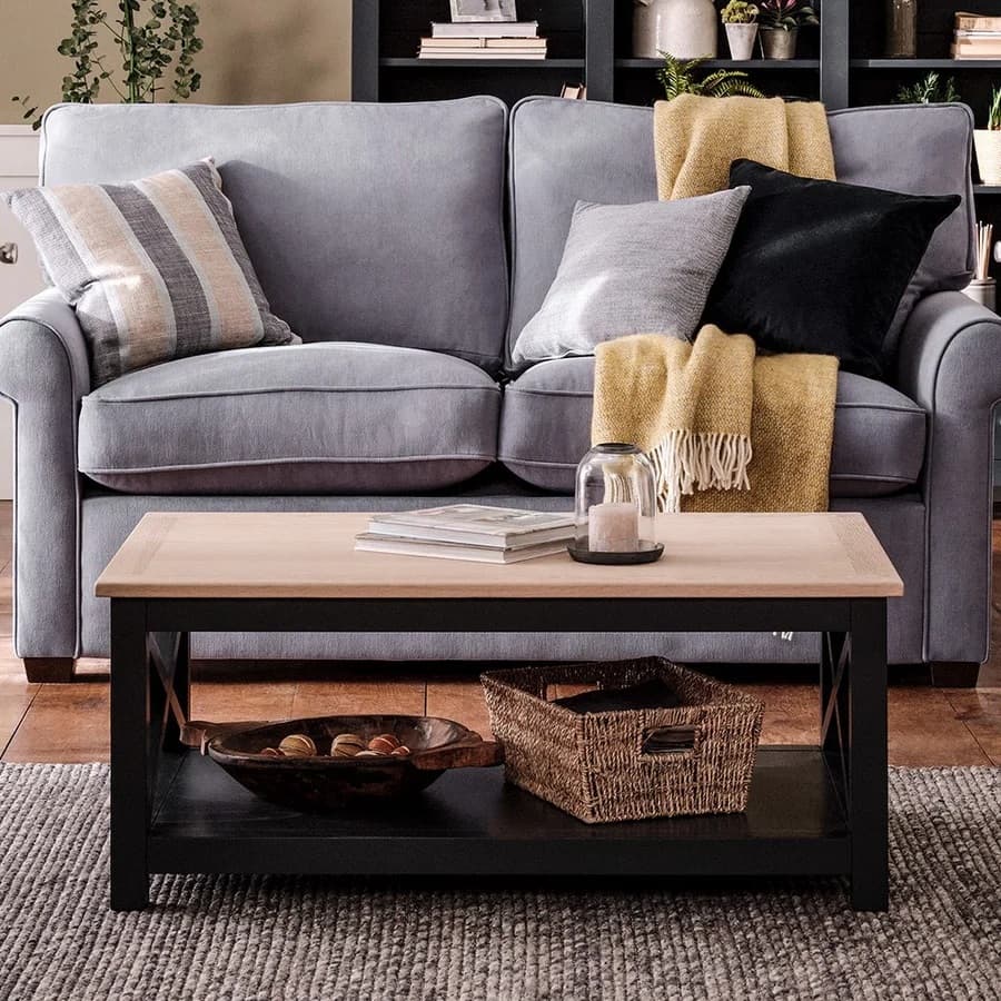 Grey sofa with pillows and a yellow throw, a wooden coffee table with books and a candle, and a shelf with plants in the background.