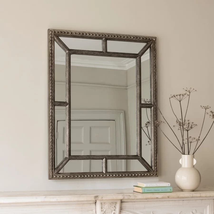 Elegant wall mirror with a decorative frame above a mantel, next to a vase with branches and two stacked books.