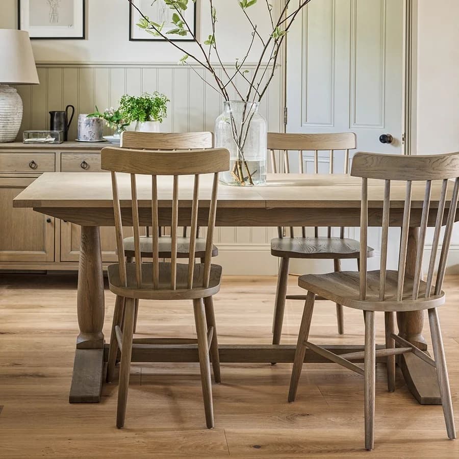 A wooden dining table with four matching chairs, a glass vase with branches, and a sideboard in a cozy, well-lit room.