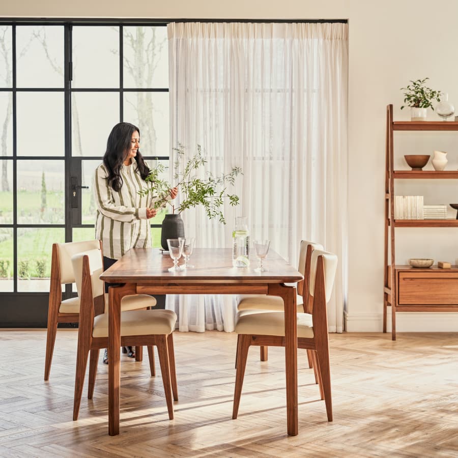 A woman arranges flowers in a vase on a wooden dining table, set in a bright room with large windows, shelves, and parquet flooring.
