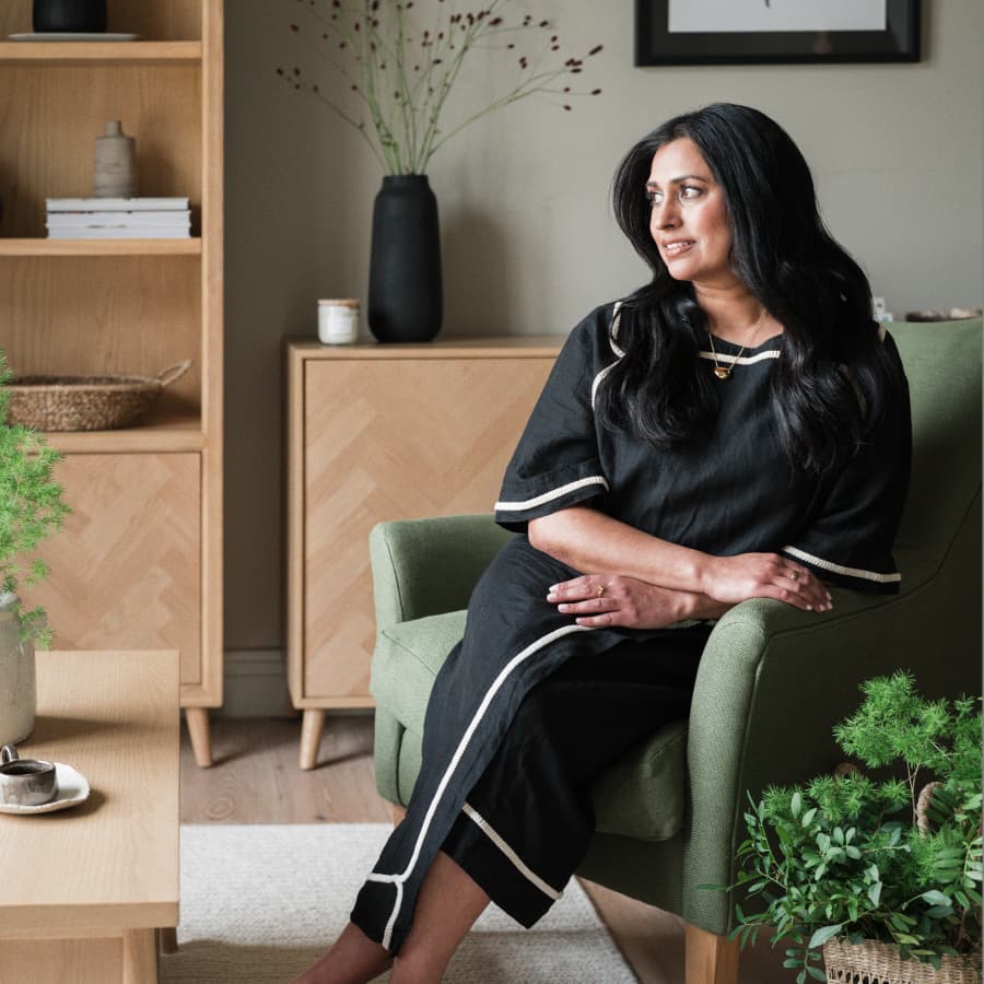 Reena Simon in a black outfit sits on a green chair in a stylish living room with plants and wooden furniture, looking to the side.