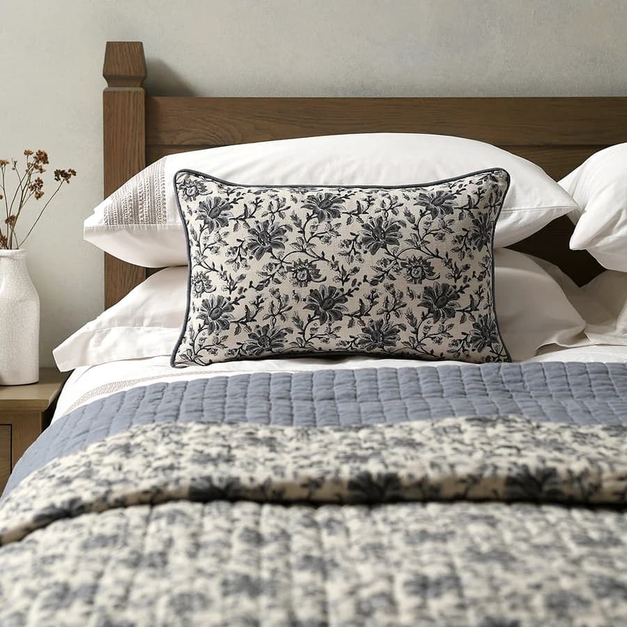 Ornate floral pillow on a bed with white pillows, wooden headboard and matching floral blanket, beside a white vase.