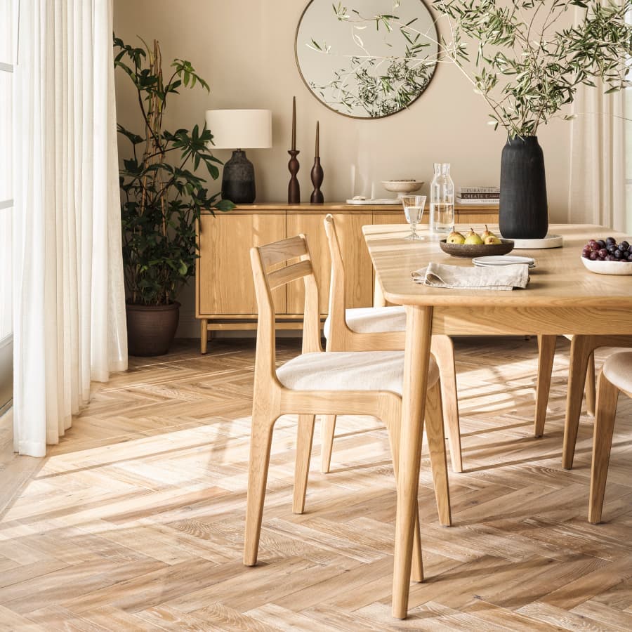 Minimalist dining room with a wooden table, light chairs, fruit bowls, and a vase. A sideboard with decor accents and plants complement the space.