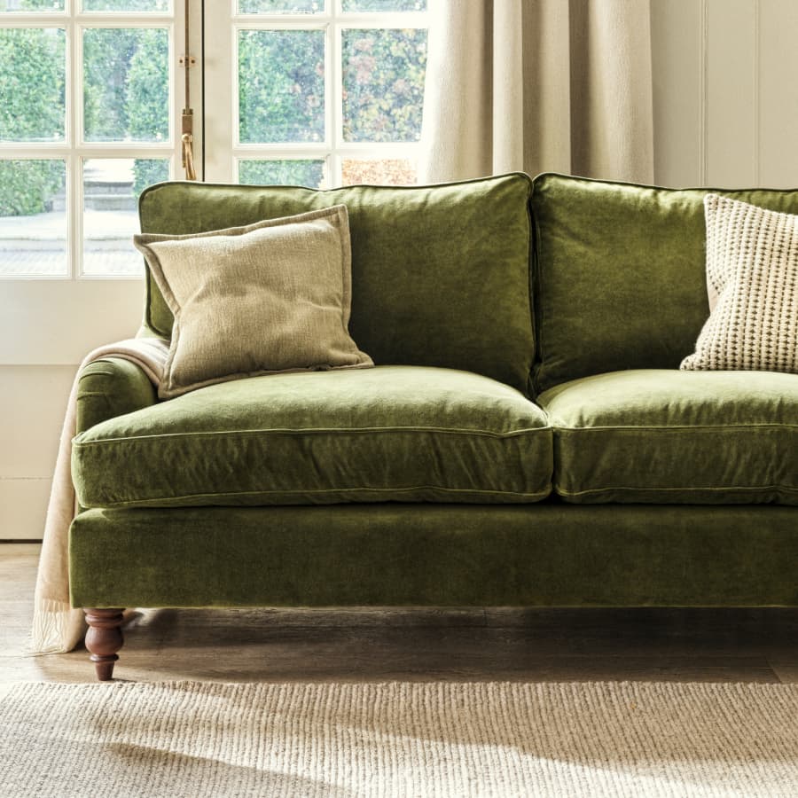 Green velvet sofa with two beige cushions in a cozy, sunlit living room near large windows and light curtains.