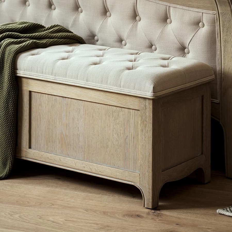 Wooden storage bench with a tufted beige cushion and a green knitted blanket draped over it, set on a wooden floor.