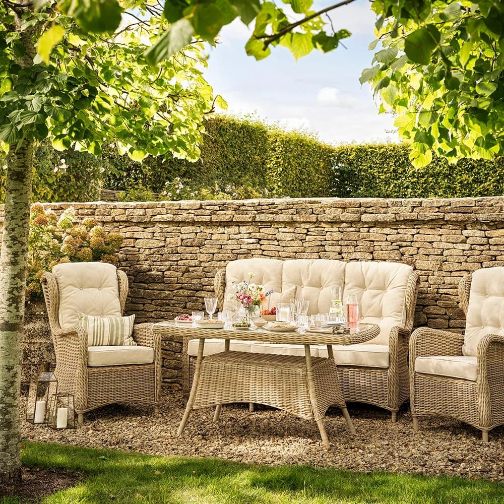 Wicker patio furniture set on a gravel area, with a stone wall, surrounded by greenery. The table is adorned with flowers and refreshments.