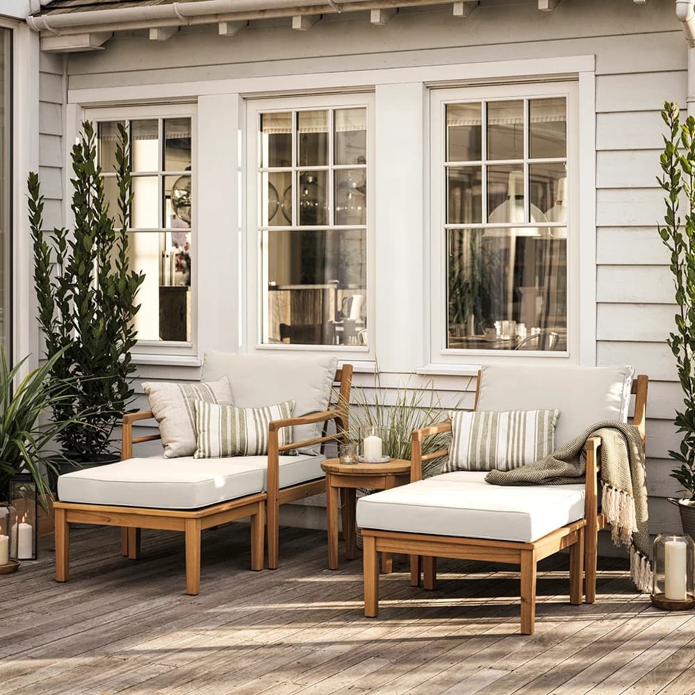 Cozy patio with wooden furniture, cream cushions, striped pillows, plant decor, and candles, set against a white-paneled house with large windows.