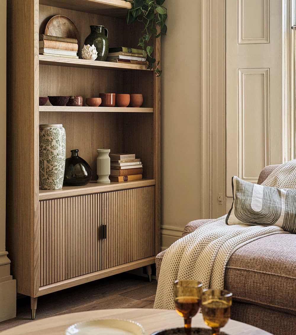 A cosy living room with a wooden bookshelf filled with books and vases, a comfortable sofa draped with a blanket, and amber glasses on a coffee table.