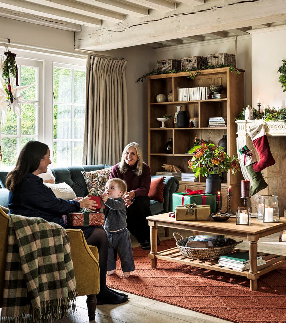 Cosy living room with two women and a child surrounded by festive decorations and wrapped gifts, next to a fireplace with stockings.