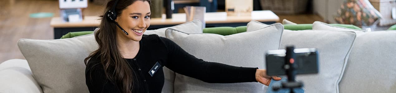 A woman with a headset sits on a neutral sofa, smiling and engaging in a video call.