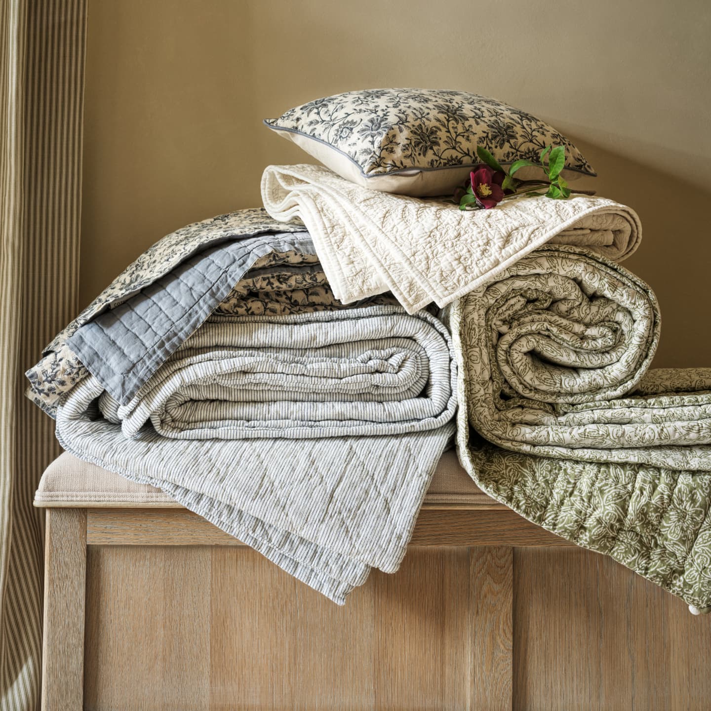 Stacked patterned quilts and a pillow with floral designs arranged on a wooden bench, accented by a purple flower.