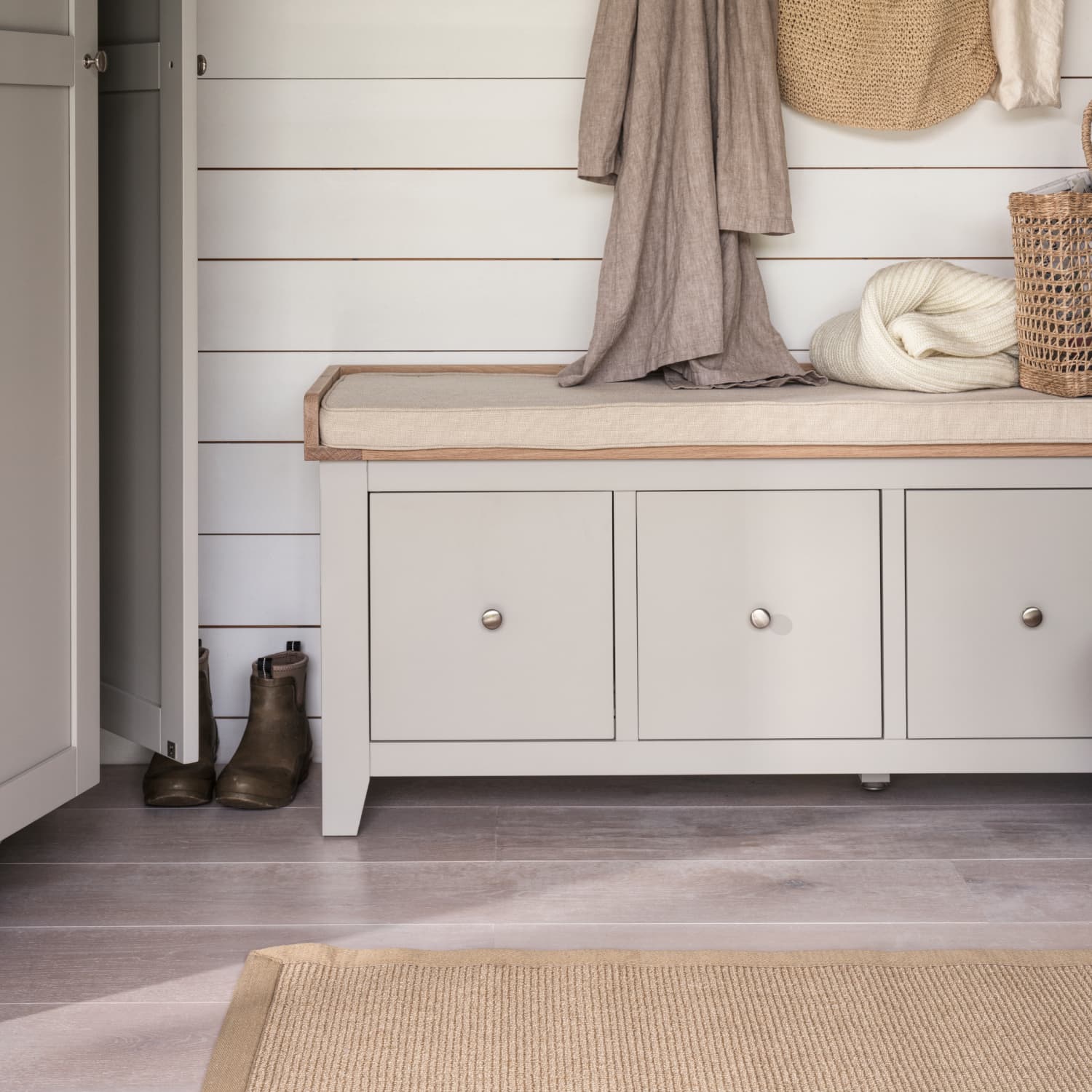 An entryway with a beige cushioned bench, boots on the floor, hanging hats and scarves, and a woven basket. Neutral tones throughout.