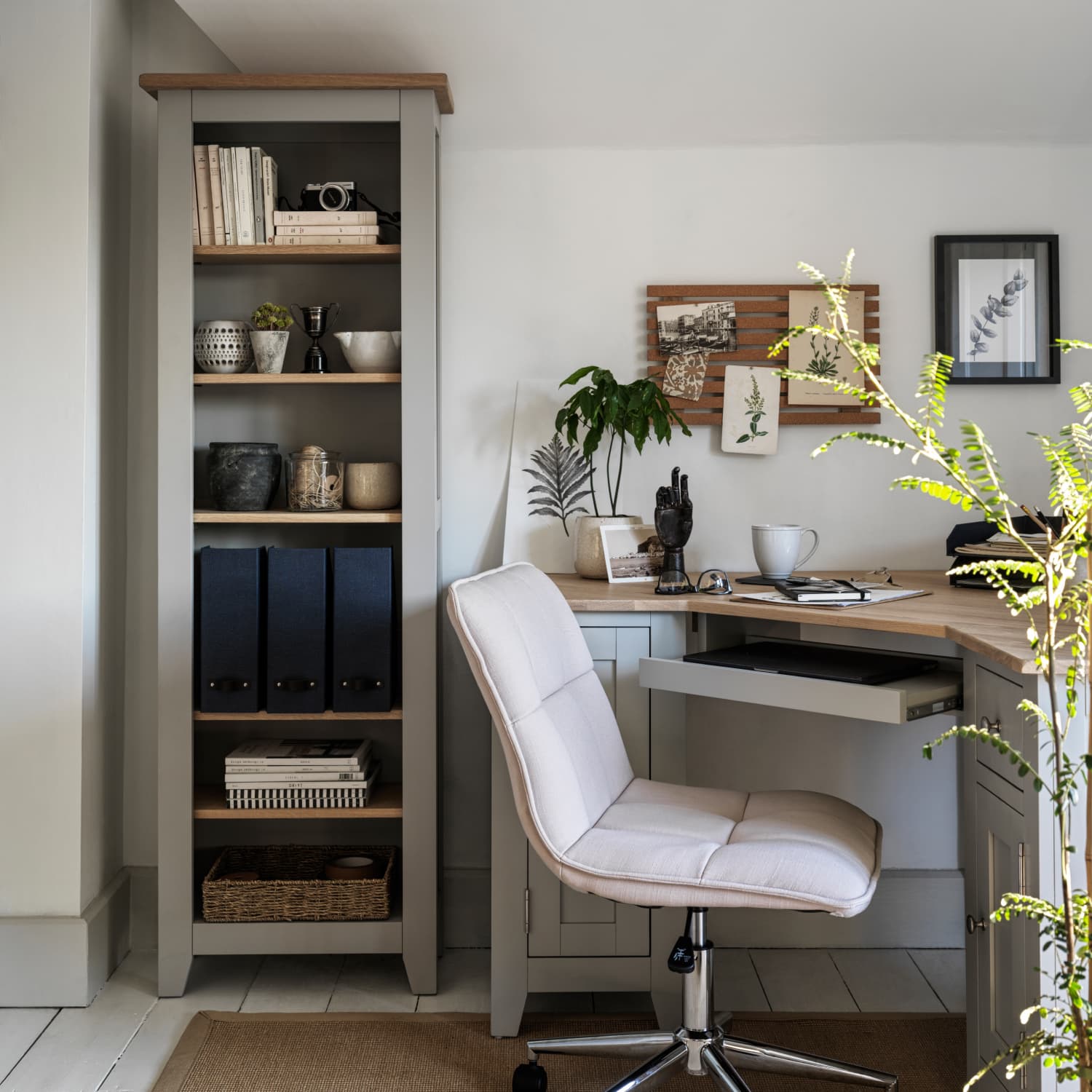 Grey home office with an upholstered chair, desk with decor and coffee cup, bookshelf with books, camera, and plants, framed art on the wall.