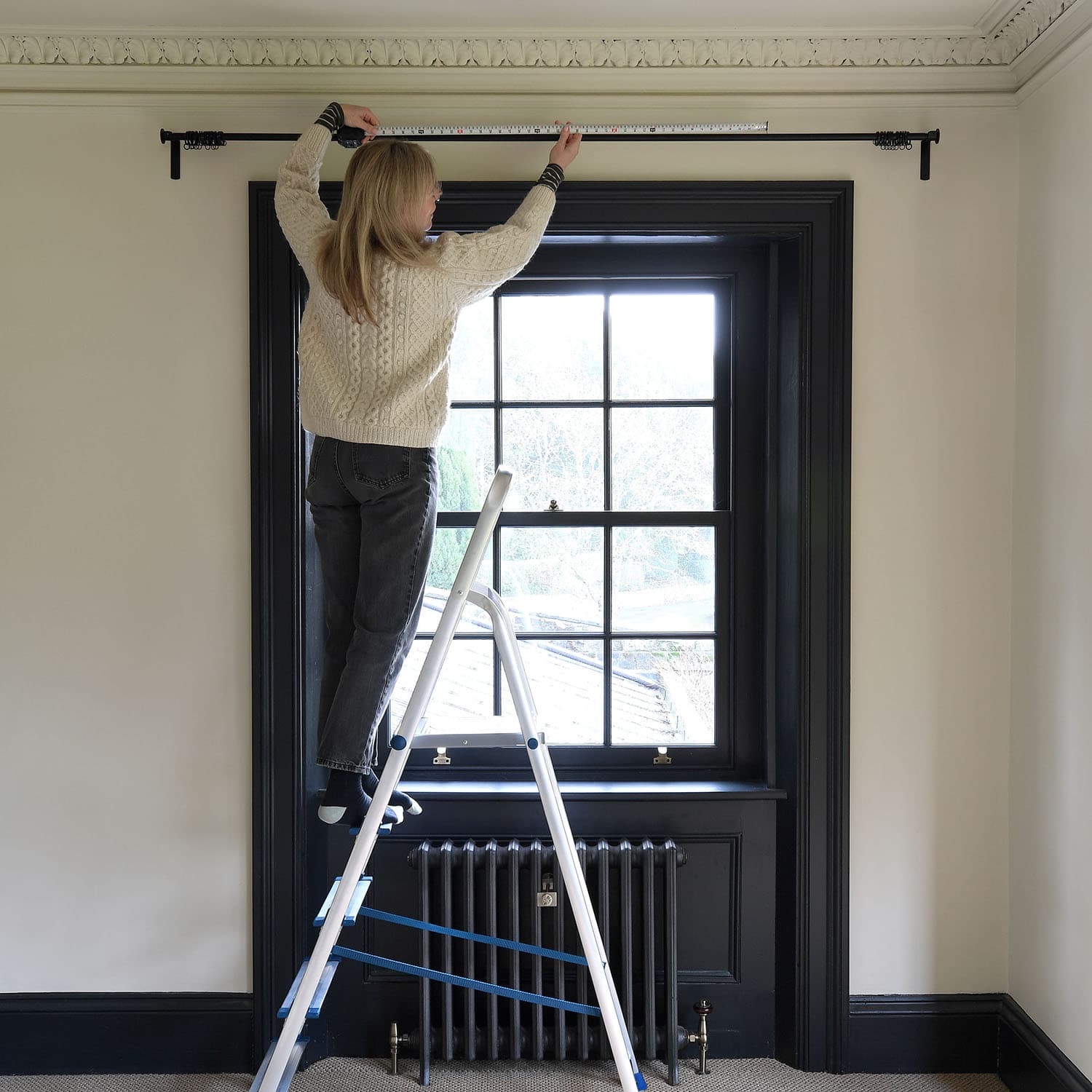 Woman standing on a ladder, measuring a curtain rod above a window with ornate molding in a room with black trim.