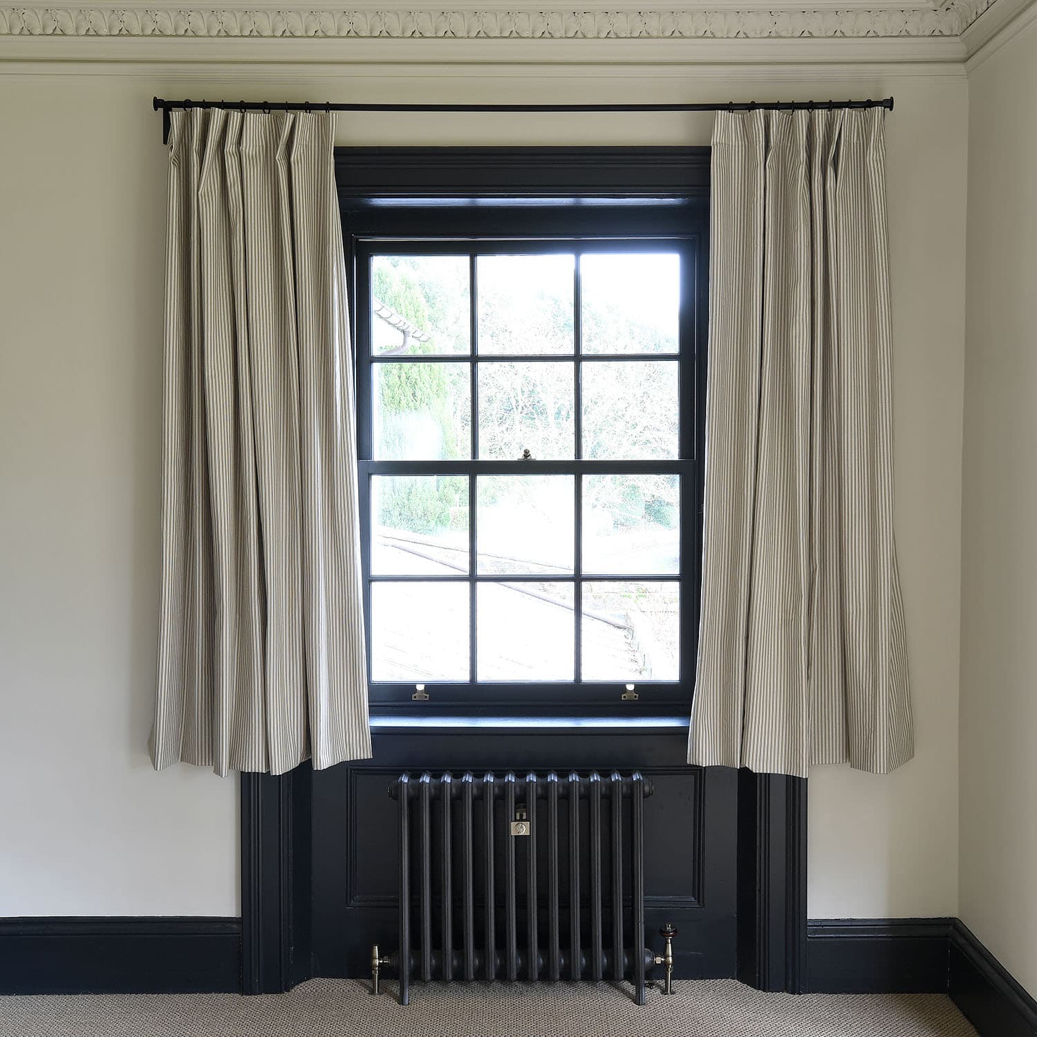 A classic window with black trim features striped curtains and an ornate radiator beneath, set against a cream wall with decorative molding.