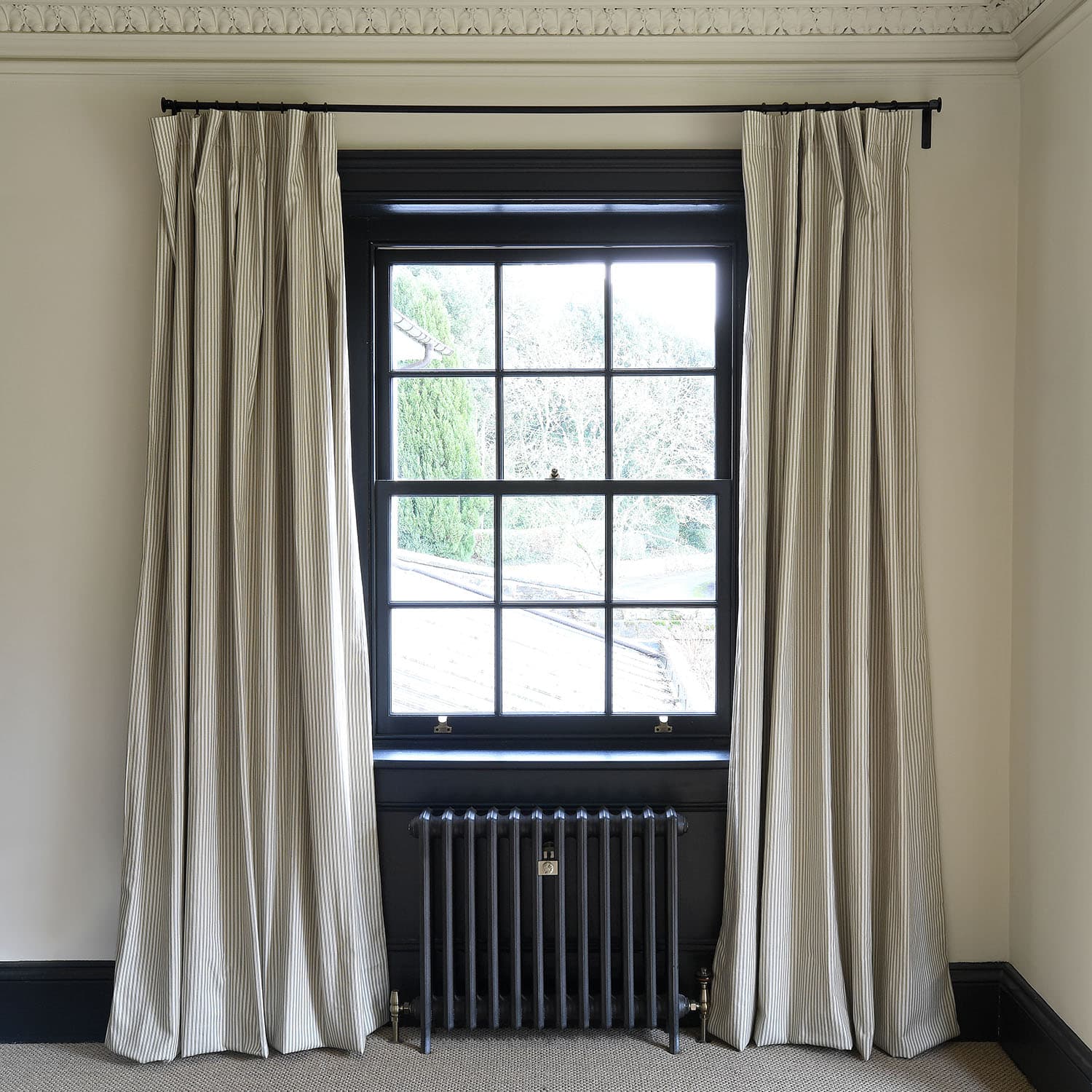Large window with black frame, covered by beige striped curtains, above a black radiator. Outside is a leafy view.