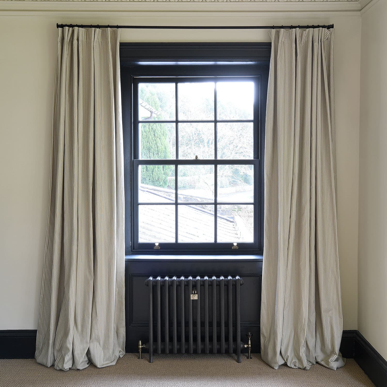 Bright window with black frame, striped curtains, and a black radiator beneath on a light-colored wall.