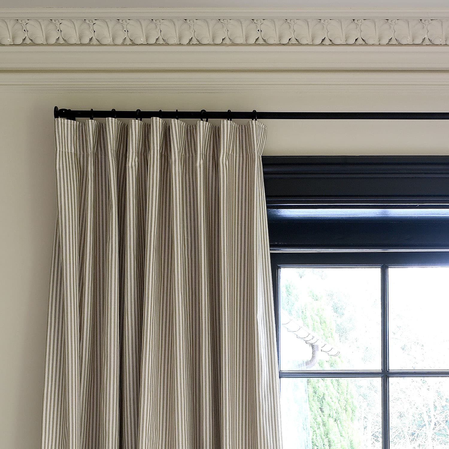 Striped beige curtains hang from a black rod, next to a black-framed window, beneath ornate white crown molding.