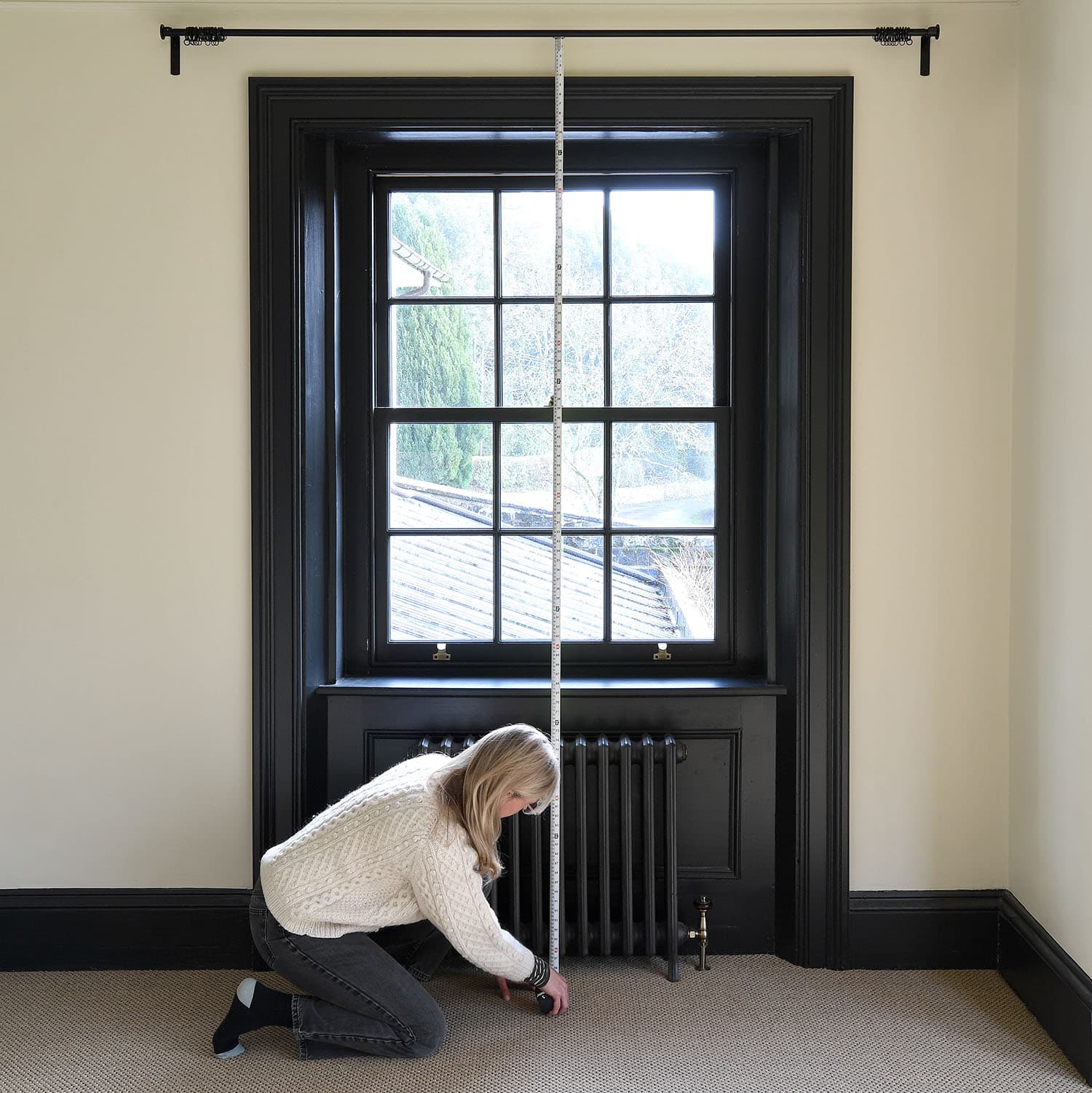 A person measures the height of a large window with a tape measure in a room with cream walls and black trim.