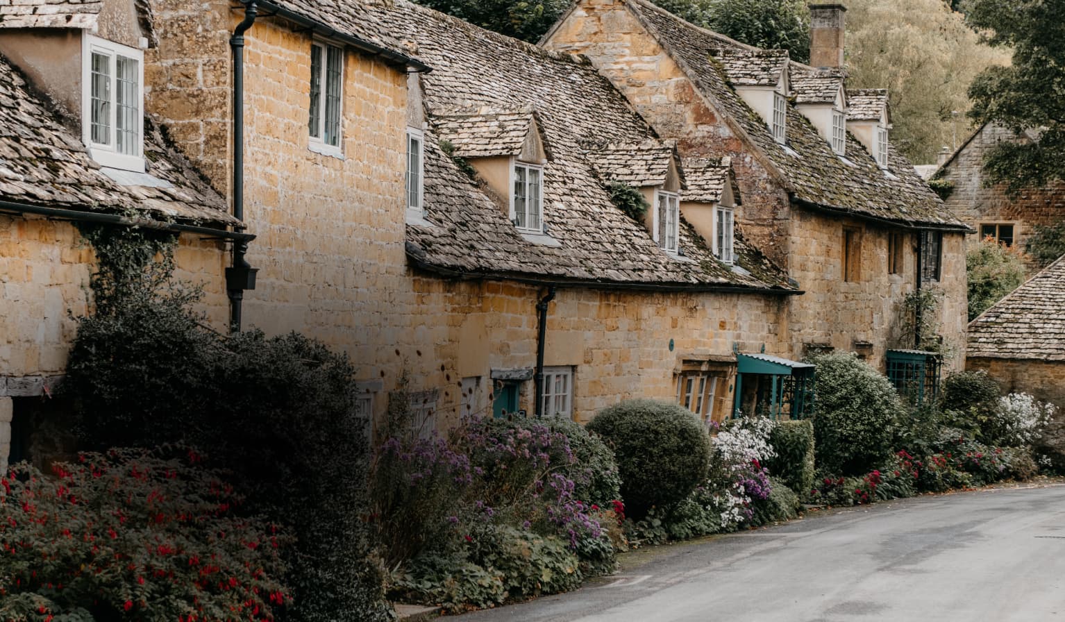 Quaint stone cottages with sloped, mossy roofs line a narrow road, surrounded by lush, colorful gardens and greenery.
