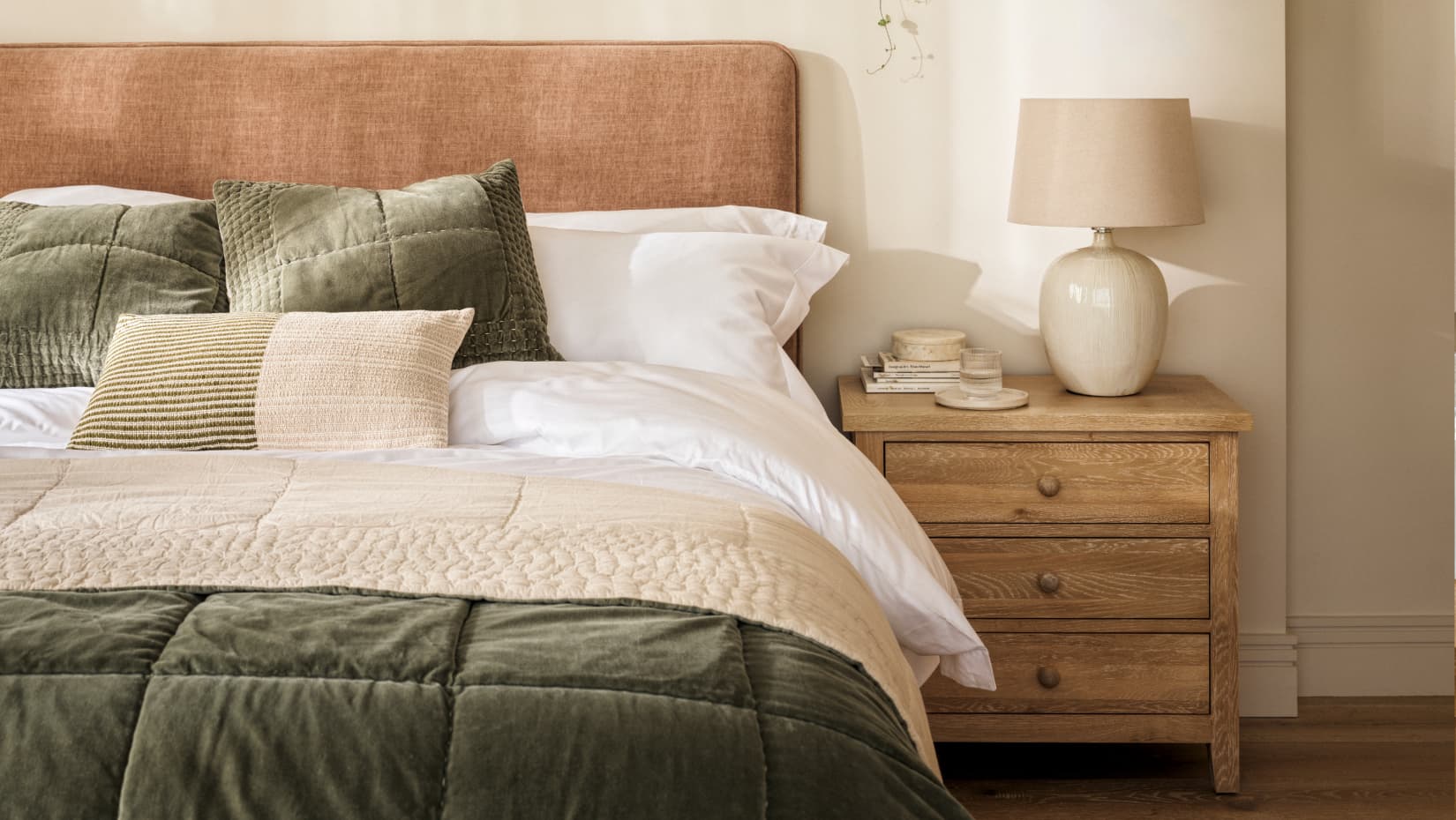 Cozy bedroom with olive green and beige bedding, plush pillows, a wooden nightstand holding books, a lamp, and candles.