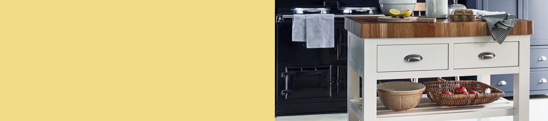 A kitchen with a wooden-topped island, drawers, and shelves, holding kitchenware and produce, against a stove and cabinetry backdrop.