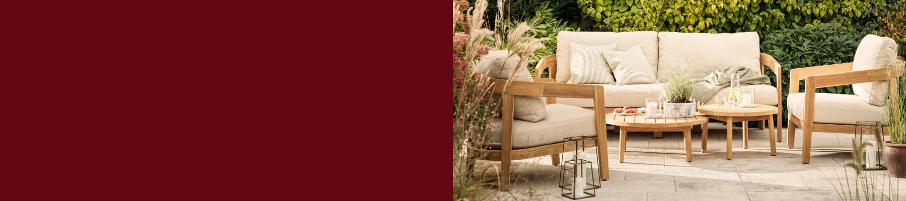 Outdoor patio with beige cushioned wooden furniture, two round tables holding candles and potted plants, surrounded by greenery and flowers.