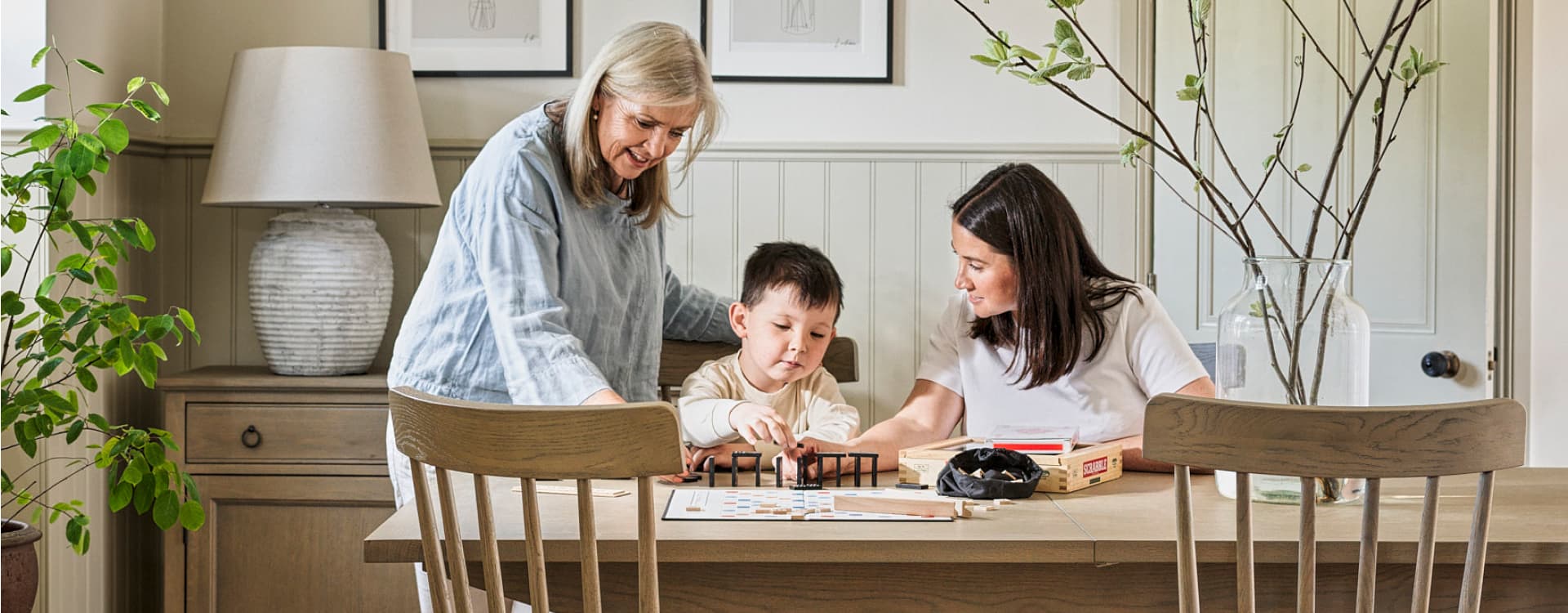 A child and two women enjoy a board game together at a wooden dining table, surrounded by plants and soft lighting.