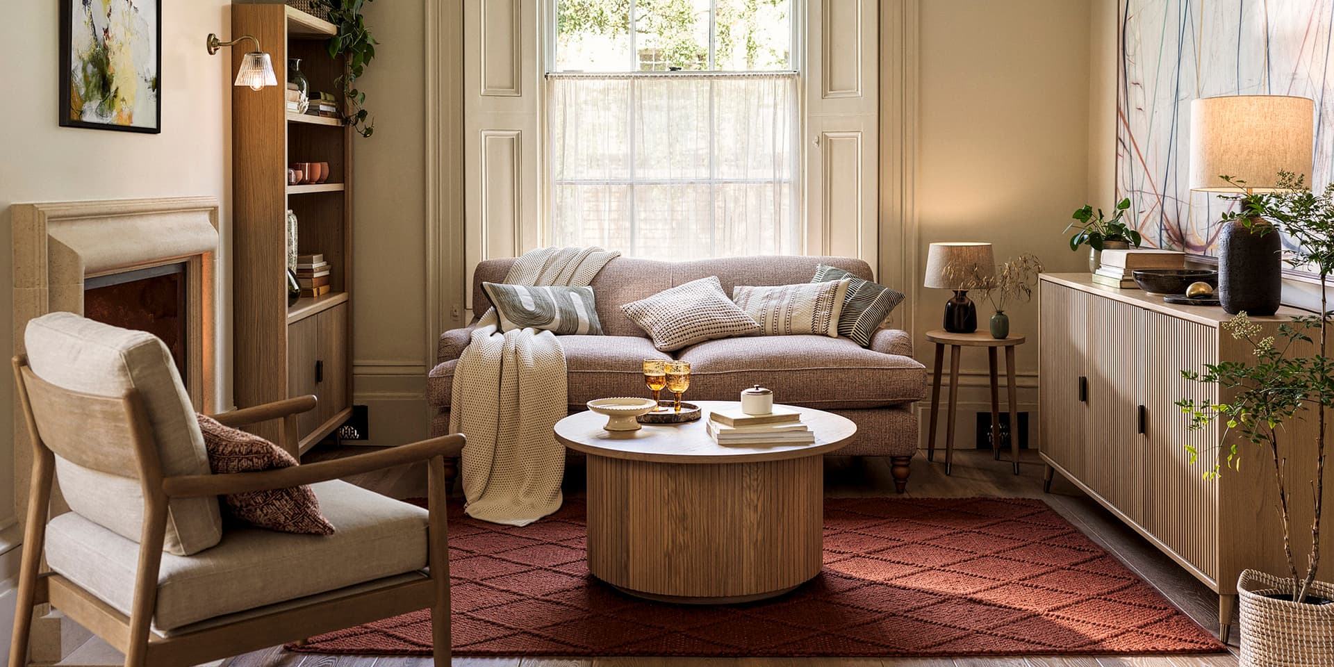 Cosy living room with a sofa, armchair, round wooden coffee table, and shelves. Soft lighting, plants, and decorative pillows enhance the ambiance.