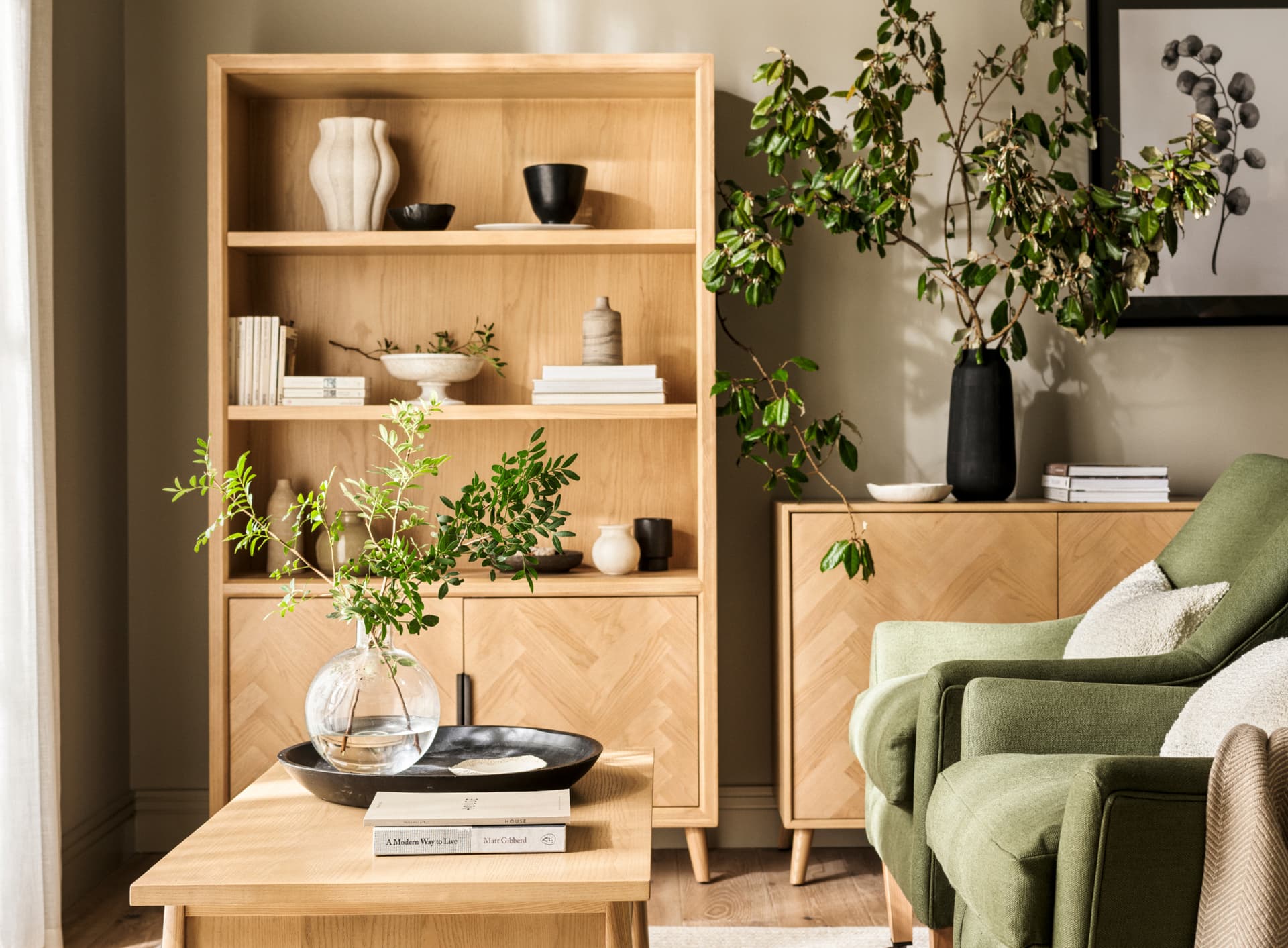 Stylish living room with a green armchair, wooden bookshelf, vases with greenery, and soft lighting.