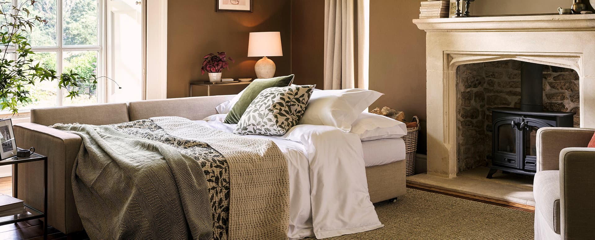 A cosy living room with a sofa bed, green cushions, a fireplace, and a window with greenery outside. Lamps and plants add warmth to the space.