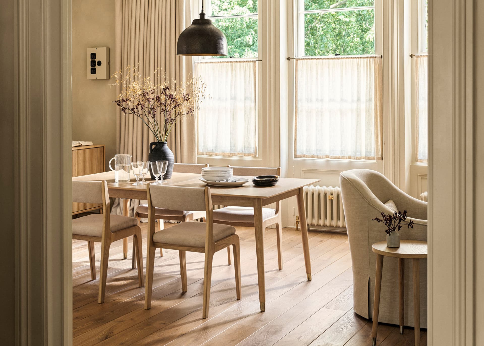 Bright dining room with wooden furniture, a table set for four, a black vase with flowers, and large windows with sheer curtains.