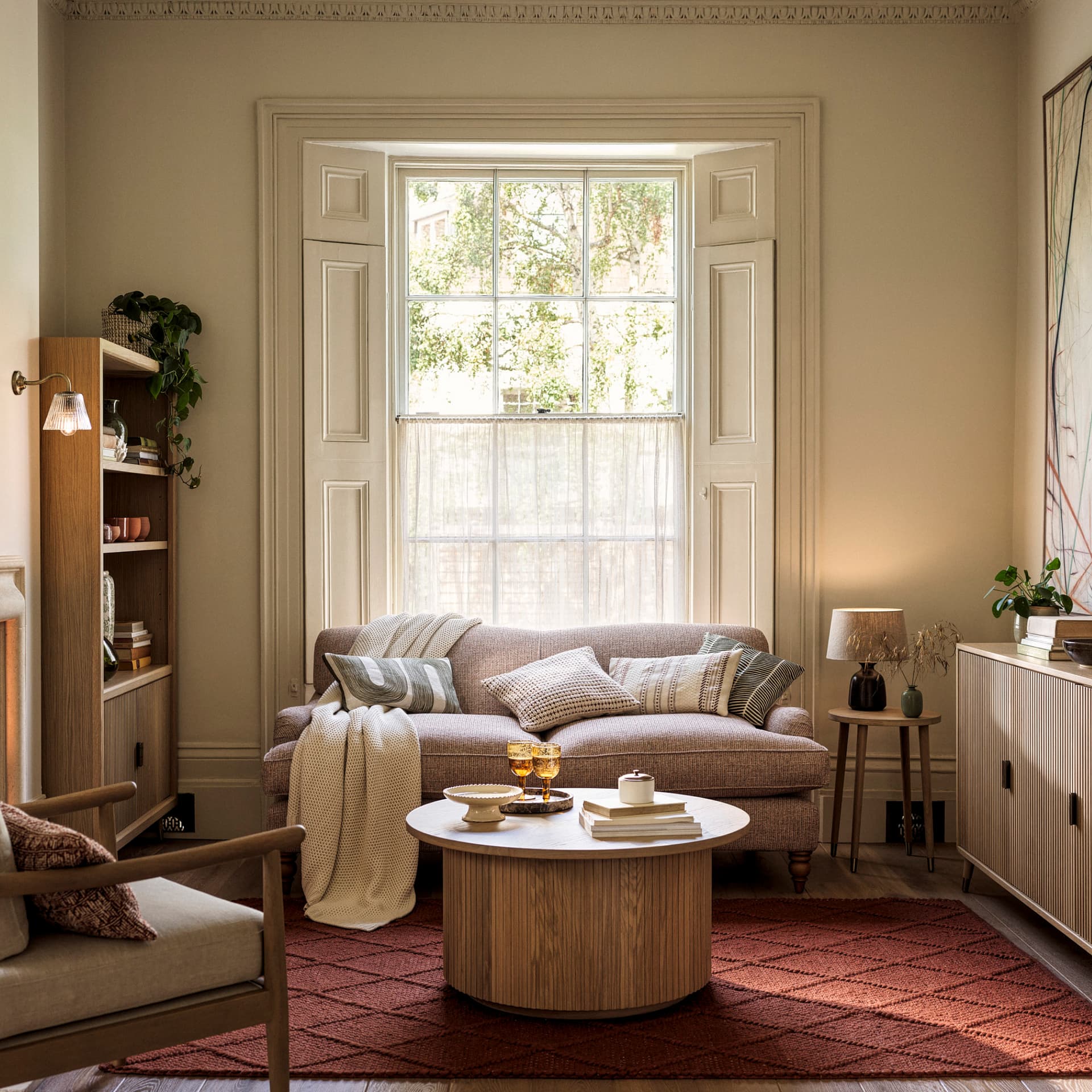 Cosy living room with a beige sofa, round wooden coffee table, white framed window, bookshelves, and a red patterned rug. Soft lighting enhances warmth.