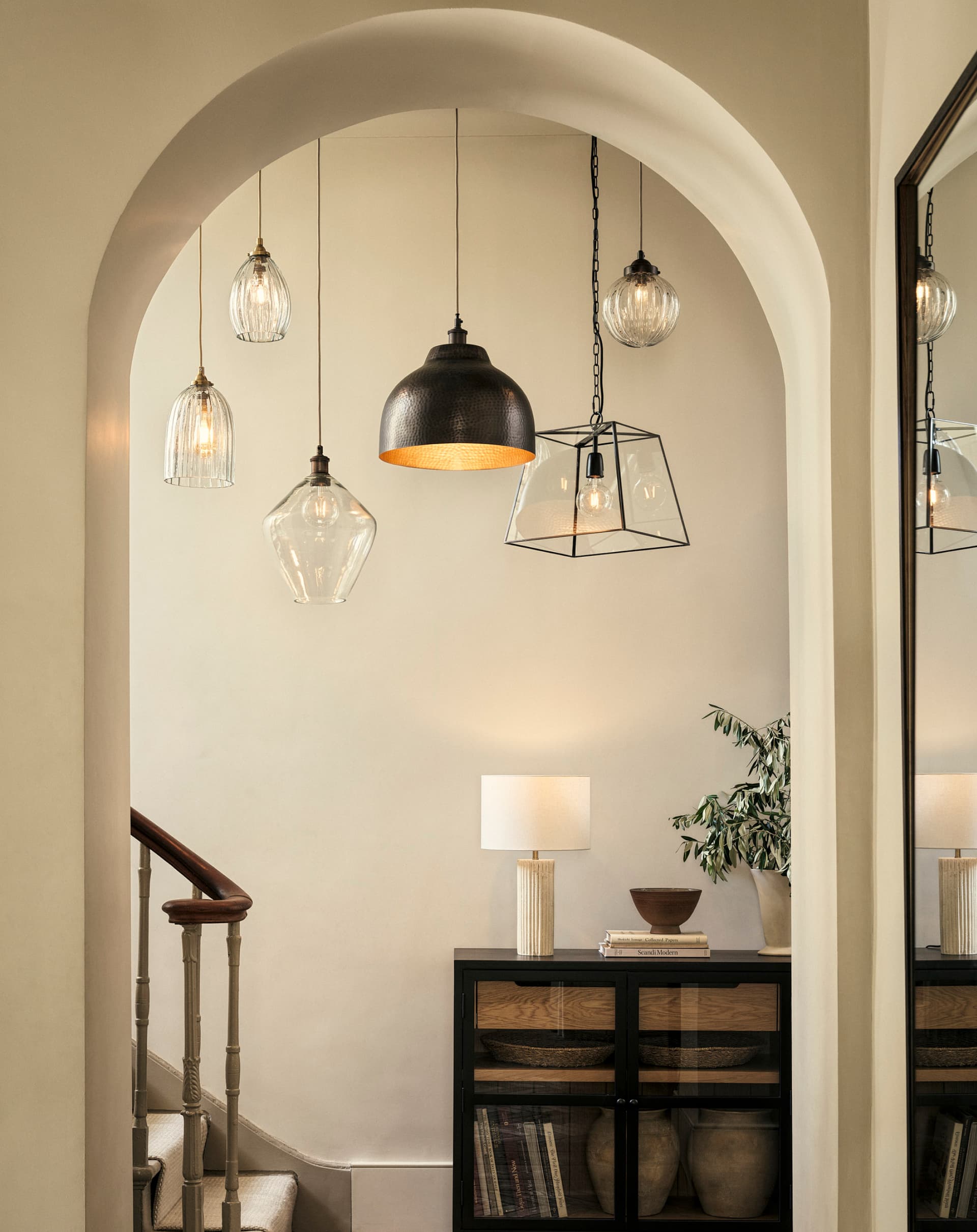 An elegant hallway features a variety of pendant lights, a staircase, a black cabinet, and a lamp with decor, reflected in a large mirror.