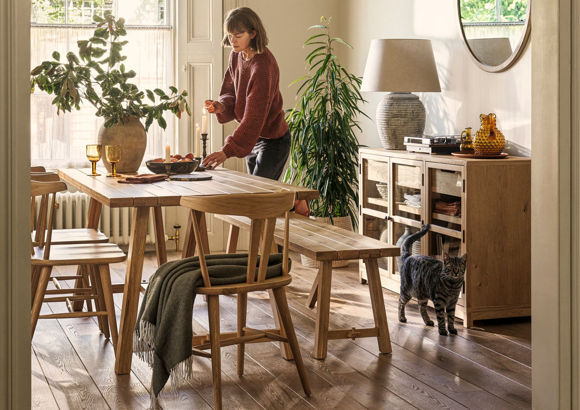 A person arranging candles on a wooden dining table in a cosy room, with plants, a cat, and warm lighting creating a homely atmosphere.