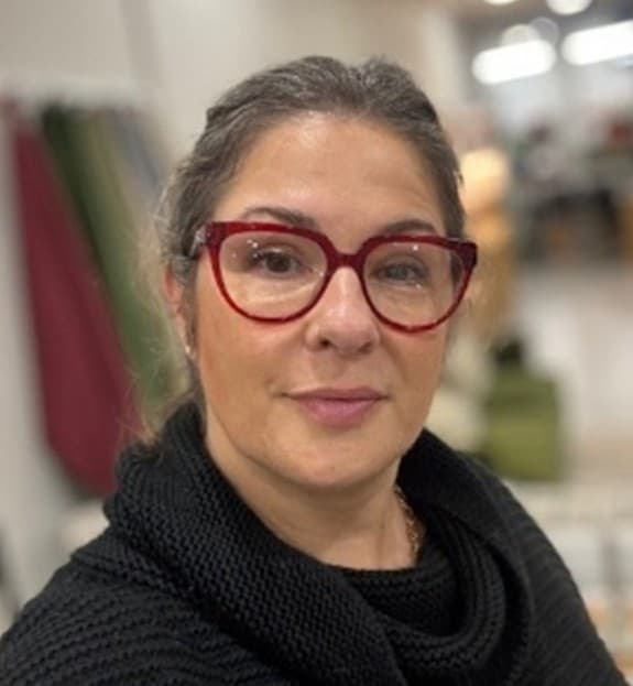 Person wearing red glasses and a black sweater, posing indoors with blurred background displaying fabrics.