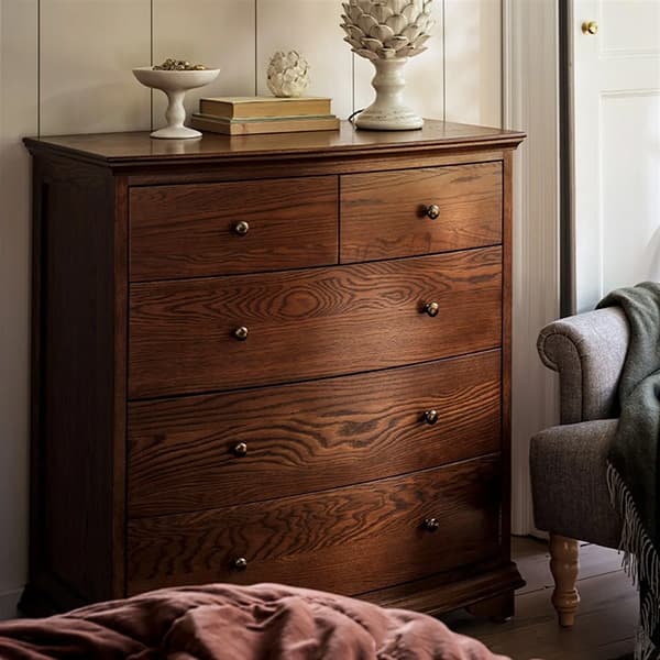 A dark wooden dresser with five drawers, adorned with decorative items, stands next to a cushioned chair with a draped blanket in a cozy room.