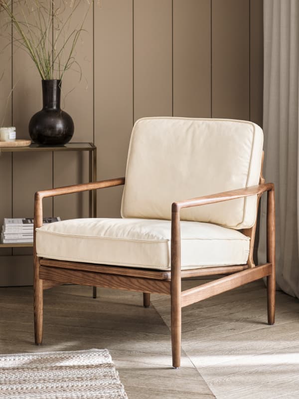 Wooden armchair with cream cushions in a cozy room, featuring a black vase on a console table and a textured rug on the floor.
