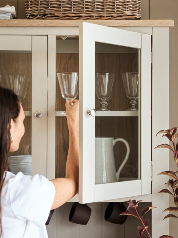 A woman reaches for a wine glass in a kitchen cabinet with glass doors. Inside are more glasses and a white pitcher. A basket sits on top.