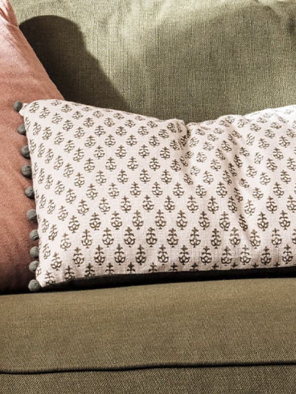 White patterned cushion with pom-pom trim on a green sofa, beside a pink pillow.