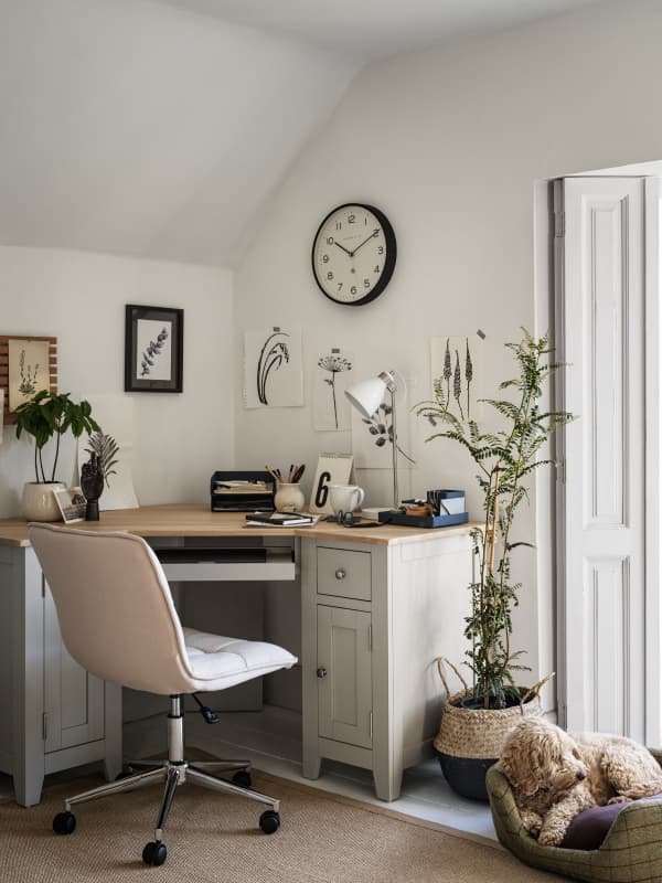 Home office with a beige chair, grey pained desk with a wooden top, plants, wall clock, and framed art. Natural light from a door; dog resting on a cushion.