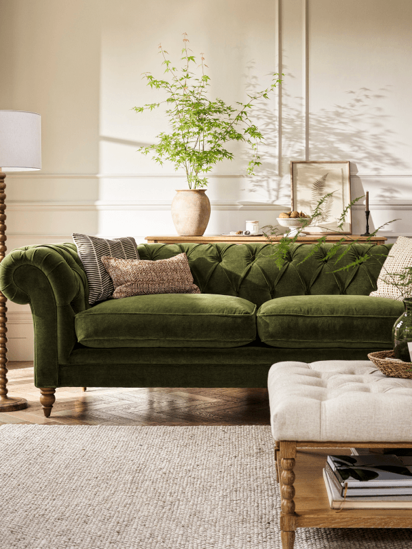 A cosy living room with a green velvet sofa, assorted pillows, a wooden coffee table, potted plants, and a large woven rug on a wooden floor.
