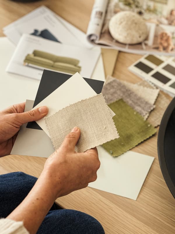 Person holding fabric swatches and paper samples over a table with various colors and textures, suggesting interior design planning.