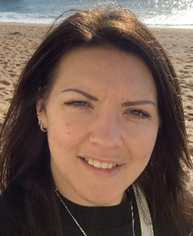 Woman smiling on a beach with the sea and sand in the background, wearing a black top and a necklace.