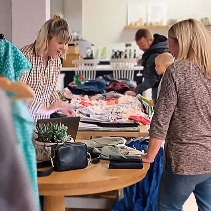 Two women looking at clothes on a table in a cozy indoor setting, with a man and child in the background near a kitchen area.