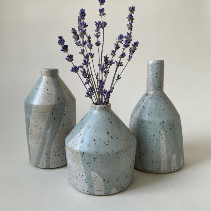 Three speckled ceramic vases, one holding sprigs of purple lavender, arranged against a neutral background.