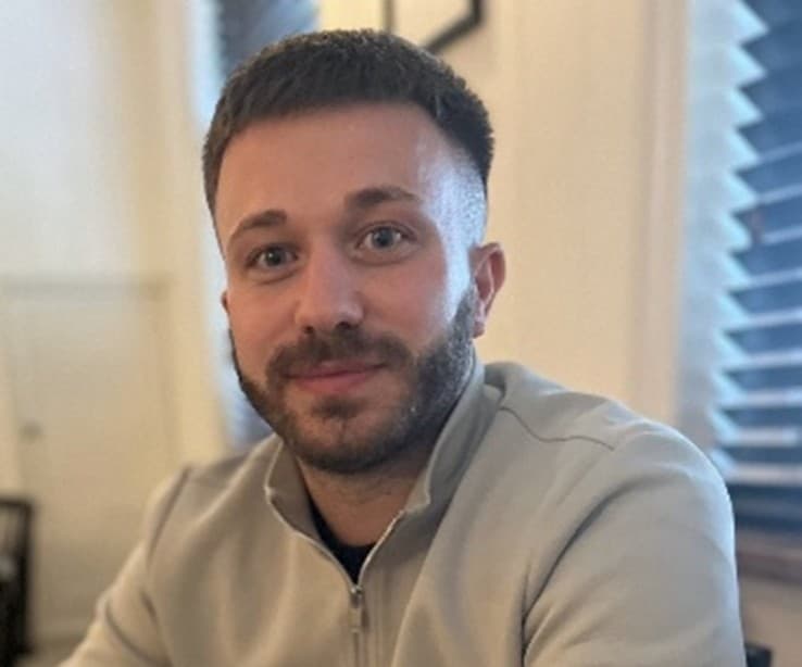 A man with short hair and a beard, wearing a light-colored jacket, smiles while sitting indoors with window blinds in the background.