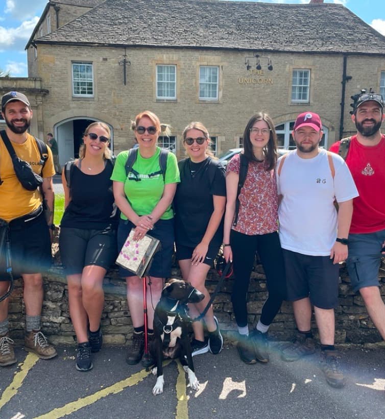 A group of seven people and a black dog pose in front of "The Unicorn" building on a sunny day, smiling at the camera.