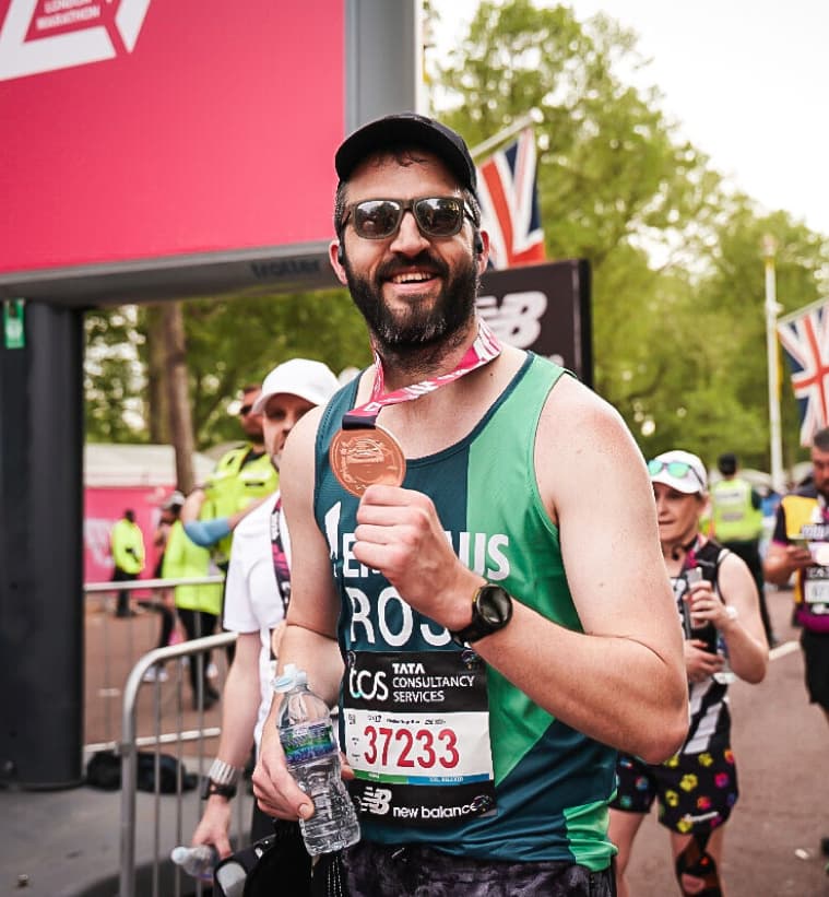 Runner with a medal smiles at the finish line of a marathon, wearing sunglasses and a cap, with a water bottle in hand.