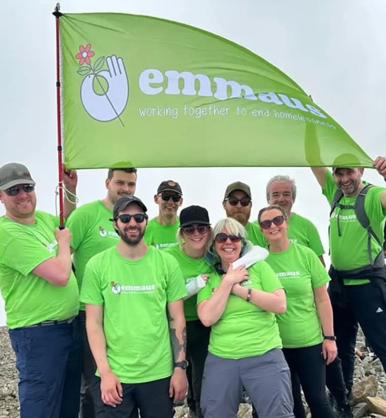 Group of people in green "Emmaus" shirts holding a banner, smiling outdoors, united for a cause against homelessness.